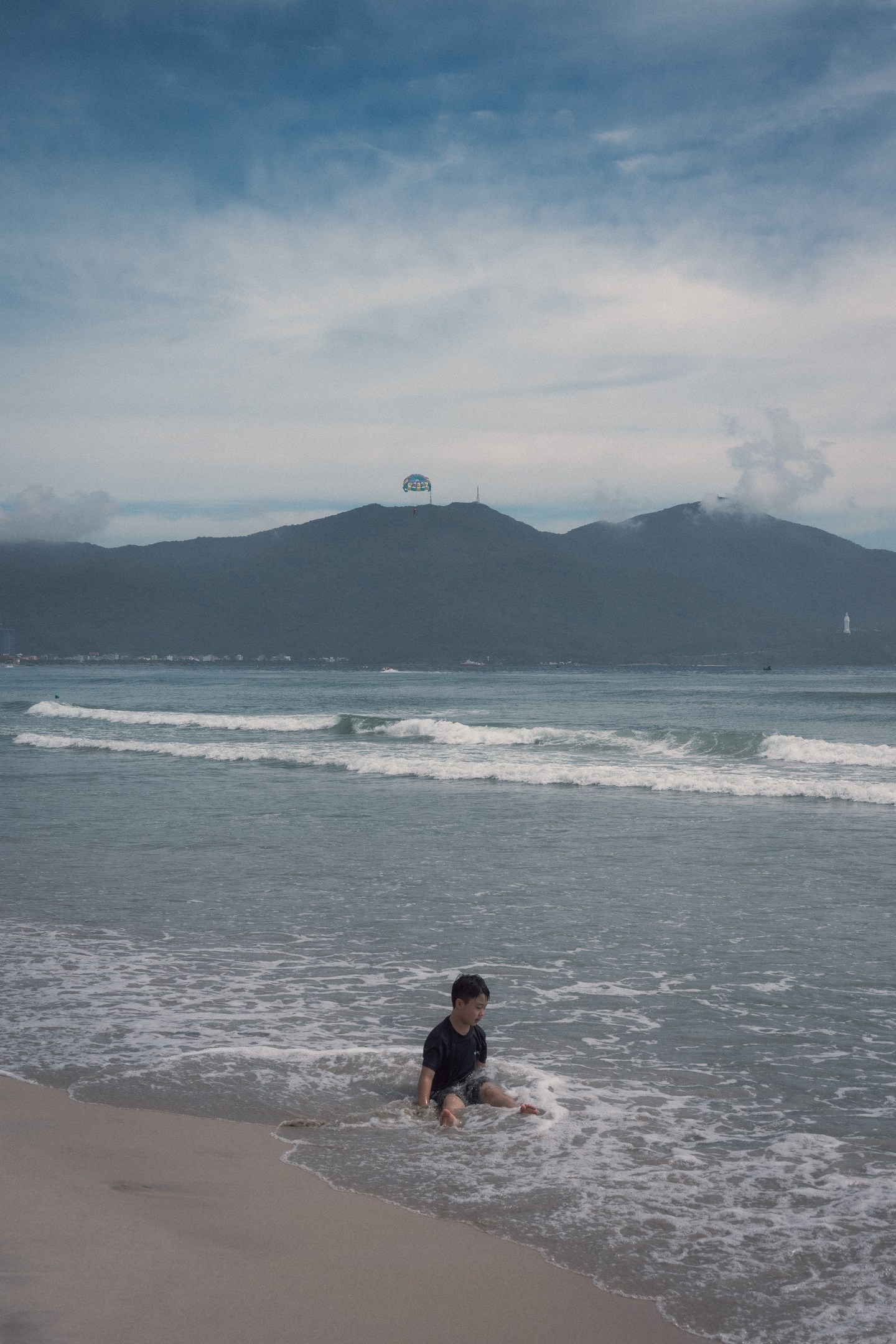 A small figure in shallow surf beneath a mountainous coastline