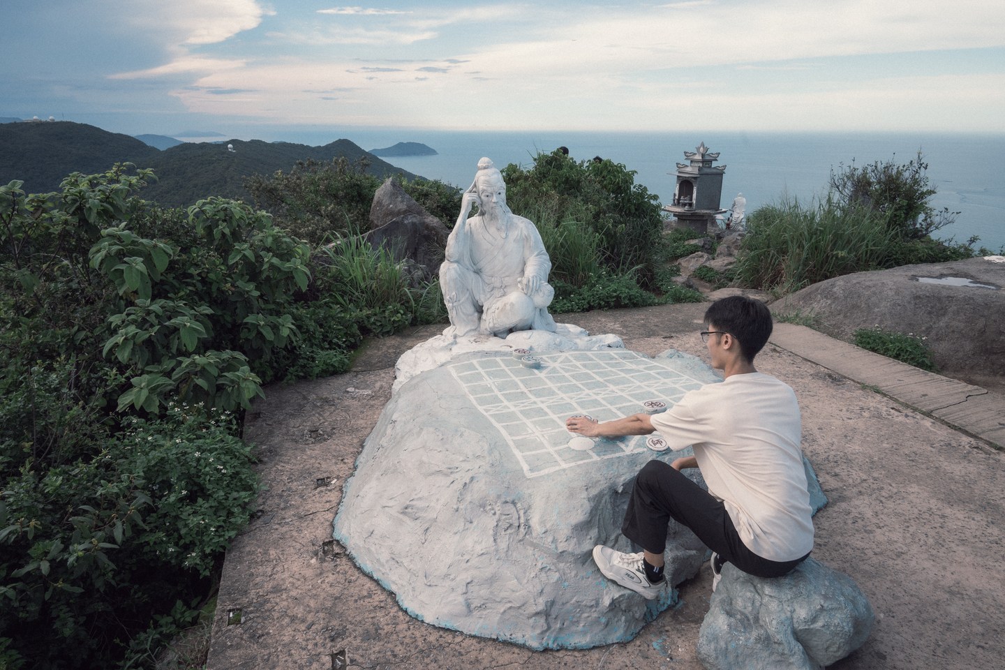 A visitor seated beside a stone sculpture on a coastal hill